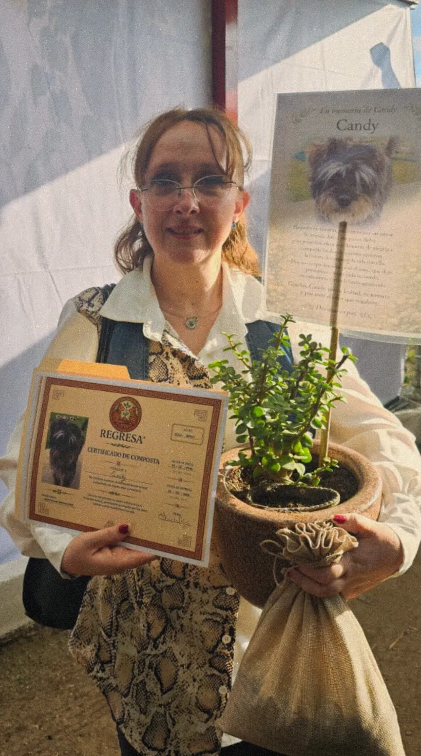 Una mujer sonríe mientras sostiene el certificado de Candy y una maceta con una planta verde y un costalito de yute, junto a la esquela en memoria de Candy
