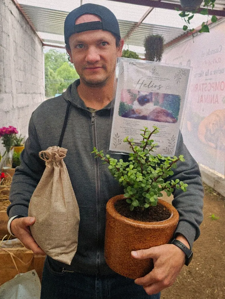 Un hombre sostiene la planta y el costalito de yute de su compañero frente a la esquela de Helios