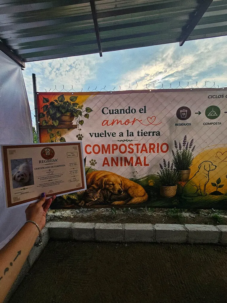 Una mano sostiene un certificado de biotransformación frente a la lona Cuando el amor vuelve a la tierra, Compostario Animal